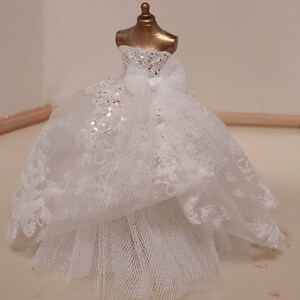 Könnte beinhalten: Ein weißes Spitzen-Hochzeitskleid mit einem Tüll-Unterrock. Das Kleid ist auf einer goldenen Puppe.
