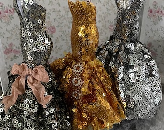 May include: Three sequined dresses hanging on a rack. The dresses are gold, silver, and black. The dresses are all very sparkly and have a lot of detail.