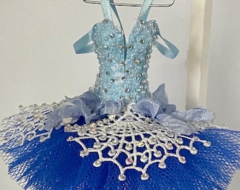 May include: A miniature ballet tutu in shades of blue, adorned with sparkling embellishments. The bodice is light blue with a V-neck, and the skirt is a darker blue tulle. The tutu hangs from a silver hanger.