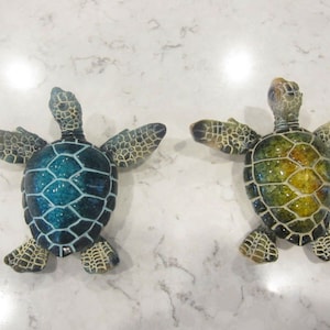 Puede incluir: Dos figuritas de tortuga marina de cerámica, una con caparazón azul y verde y la otra con caparazón verde y amarillo.
