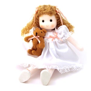 May include: A doll with curly blonde hair wearing a white dress with pink accents and holding a brown teddy bear.