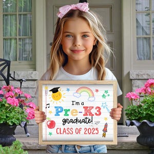 K&ouml;nnte beinhalten: Ein M&auml;dchen h&auml;lt ein Schild mit der Aufschrift "Ich bin ein Pre-K3 Absolvent! Klasse von 2023". Das Schild ist mit einem Regenbogen, Sternen, einer Rakete und einer Sonne mit einem Abschluss-Hut verziert.