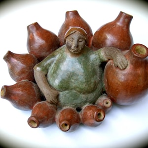 May include: A ceramic sculpture of a woman sitting on a pile of brown pottery jars. The woman is wearing a green dress and has a brown face.