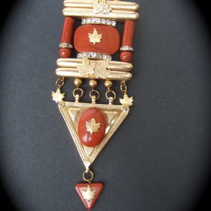 May include: A gold-tone pendant with a red stone and a gold leaf design. The pendant is attached to a gold chain with a triangular pendant at the bottom.