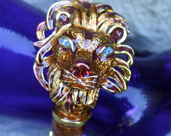 Könnte beinhalten: Eine goldfarbene Löwenkopf-Brosche mit roten und goldenen Emaildetails. Der Löwe hat blaue Augen und eine rubinrote Nase.