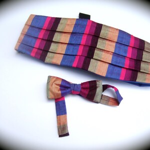 May include: A pleated cummerbund and a matching bow tie featuring a vertical striped pattern in shades of blue, magenta, dark purple, and tan. The fabric has a subtle sheen, and the set includes an adjustable strap for the bow tie.