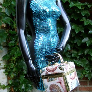 K&ouml;nnte beinhalten: Eine blaue Mosaik-Torso-Schaufensterpuppe h&auml;lt eine kleine, quadratische Handtasche im Vintage-Stil mit Muschelmuster.