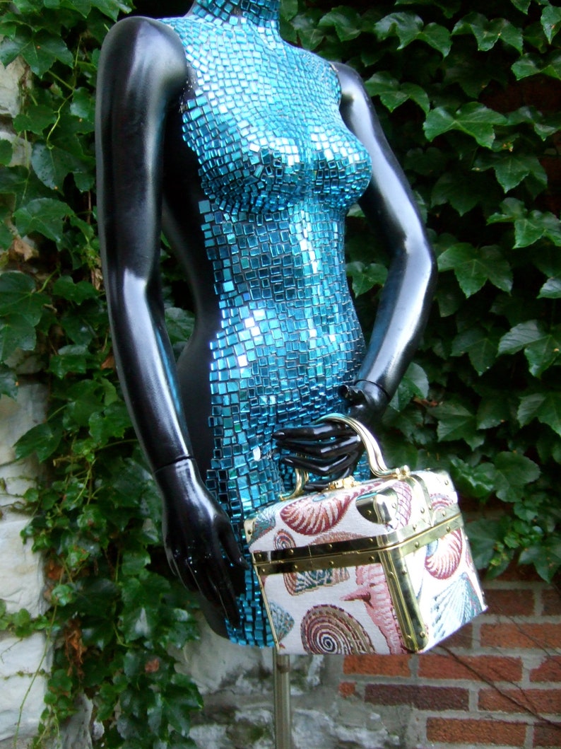 K&ouml;nnte beinhalten: Eine blaue Mosaik-Torso-Schaufensterpuppe h&auml;lt eine kleine, quadratische Handtasche im Vintage-Stil mit Muschelmuster.