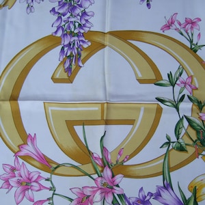 May include: A white silk scarf with a gold Gucci logo and floral designs. The flowers are pink, purple, and yellow.