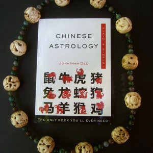 Może przedstawiać: Książka zatytułowana "Chińska astrologia: prosto i jasno" autorstwa Jonathana Dee, przedstawiająca czerwoną i białą ilustrację 12 chińskich znak&oacute;w zodiaku. Książka jest otoczona naszyjnikiem z rzeźbionych koralik&oacute;w z kości z zielonymi koralikami z jadeitu.