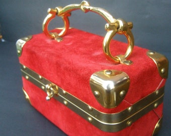 May include: A red suede box with gold metal accents and a gold handle. The box has a hinged lid and a latch closure. The box is decorated with gold studs and rivets.