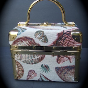 K&ouml;nnte beinhalten: Eine kleine, rechteckige, cremefarbene Handtasche mit einem goldfarbenen Henkel und Besatz. Die Handtasche hat einen tapisserieartigen Stoff mit einem Muster aus Muscheln und anderen nautischen Motiven.