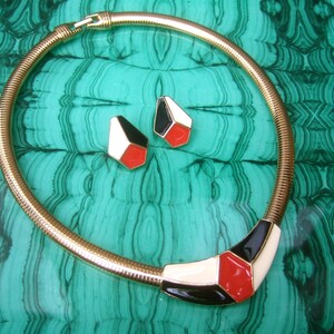 May include: A gold chain necklace with a geometric pendant featuring black, white, and red enamel. The pendant is shaped like a triangle with rounded corners. The necklace is displayed on a green and black patterned surface.