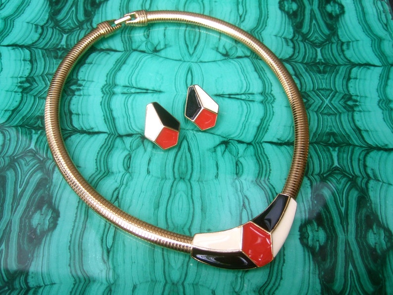 May include: A gold chain necklace with a geometric pendant featuring black, white, and red enamel. The pendant is shaped like a triangle with rounded corners. The necklace is displayed on a green and black patterned surface.