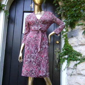 May include: A patterned wrap dress in shades of burgundy, white, and black is displayed on a mannequin. The dress features a V-neckline, long sleeves, and a matching belt. The mannequin is positioned in front of a black door.