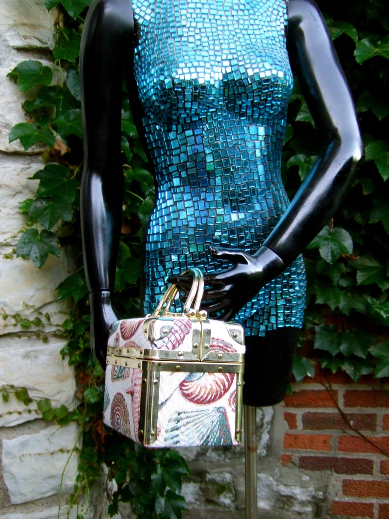 K&ouml;nnte beinhalten: Ein blaues Mosaikkleid mit einer gold-wei&szlig; gemusterten Box-Handtasche. Die Handtasche hat einen Klappdeckel und einen Messingverschluss.