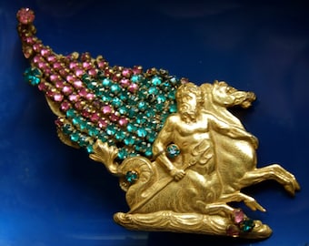 May include: A gold-toned brooch depicting a mythical creature with wings made of pink and green rhinestones. The creature is riding a horse and holding a trident.