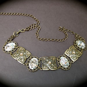 May include: A gold chain belt with a decorative center piece featuring five oval-shaped clear rhinestones set in ornate metal frames. The chain is made of small, linked circles.