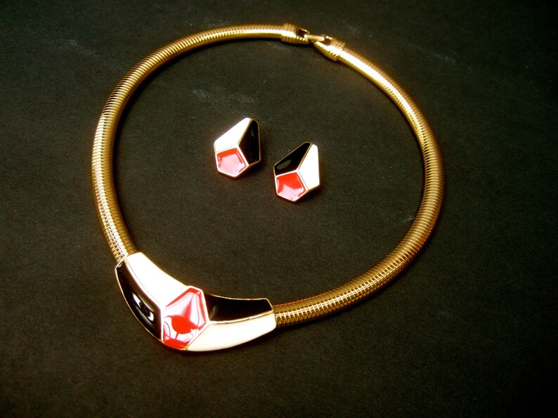 May include: A gold chain necklace with a black, white, and red geometric pendant. The necklace comes with a pair of matching earrings.