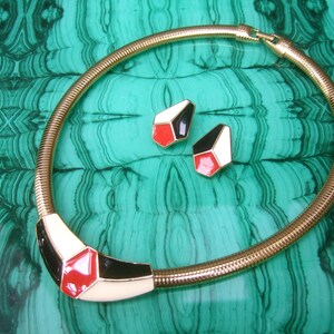 May include: A gold snake chain necklace with a geometric pendant featuring red, white, and black enamel. The pendant is shaped like a hexagon with rounded corners. The necklace is paired with matching earrings.