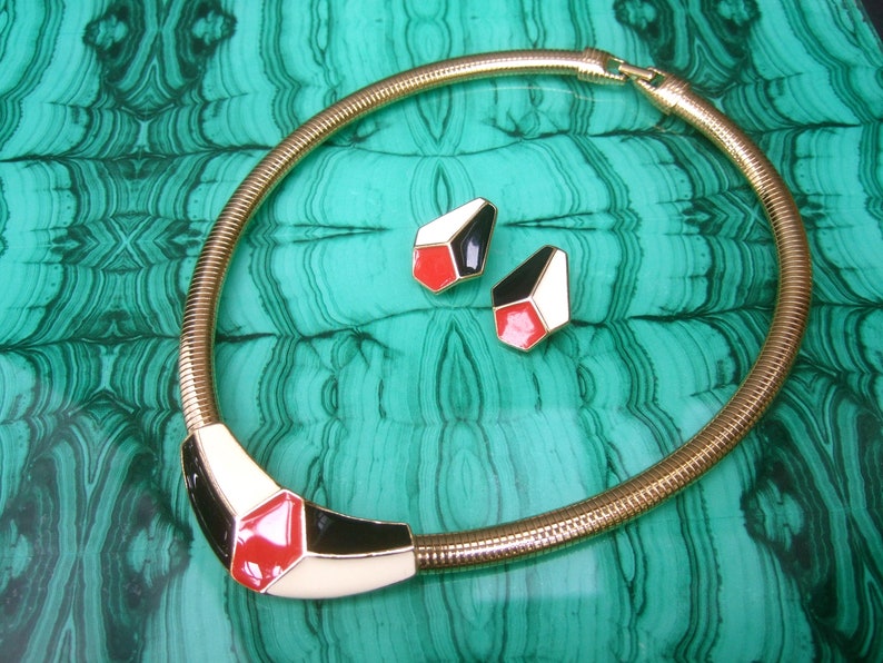 May include: A gold snake chain necklace with a geometric pendant featuring red, white, and black enamel. The pendant is shaped like a hexagon with rounded corners. The necklace is paired with matching earrings.