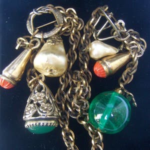 May include: A gold chain necklace with multiple charms, including a pearlescent white charm, a green glass bead, and a red and gold charm. The necklace has a clasp closure.