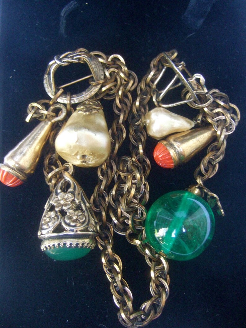 May include: A gold chain necklace with multiple charms, including a pearlescent white charm, a green glass bead, and a red and gold charm. The necklace has a clasp closure.