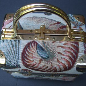 K&ouml;nnte beinhalten: Eine kleine, quadratische, wei&szlig;e und goldene Handtasche mit einem Tapisserie-Design aus Muscheln und floralen Mustern. Die Handtasche hat einen goldenen Griff und Messingakzente.