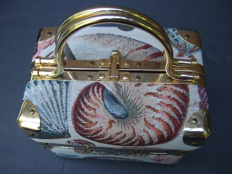 K&ouml;nnte beinhalten: Eine kleine, quadratische, wei&szlig;e und goldene Handtasche mit einem Tapisserie-Design aus Muscheln und floralen Mustern. Die Handtasche hat einen goldenen Griff und Messingakzente.