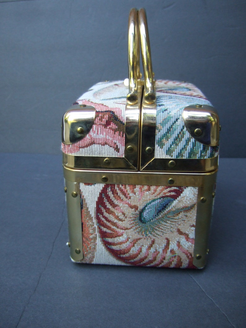 K&ouml;nnte beinhalten: Eine vintage, goldfarbene, rechteckige Handtasche mit einem Tapisserie-Stoffbezug, der ein Muschelmotiv in Rosa-, Blau- und Gr&uuml;nt&ouml;nen zeigt. Die Tasche hat einen oberen Griff und einen Verschluss aus Messing.