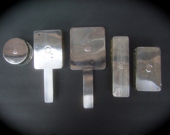 May include: A set of five silver-toned vanity items, including a round compact, two rectangular brushes, a square brush, and a rectangular box. All items have a ridged design.