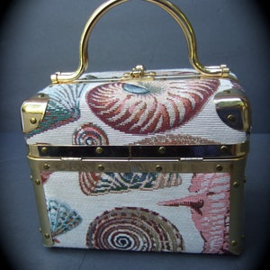 K&ouml;nnte beinhalten: Eine kleine, rechteckige Handtasche mit goldenem Besatz und einem oberen Henkel. Die Handtasche ist mit einem wei&szlig;en Stoff mit einem bunten Muschelmuster bedeckt.