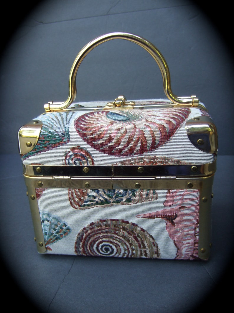 K&ouml;nnte beinhalten: Eine kleine, rechteckige Handtasche mit goldenem Besatz und einem oberen Henkel. Die Handtasche ist mit einem wei&szlig;en Stoff mit einem bunten Muschelmuster bedeckt.