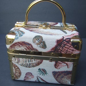 K&ouml;nnte beinhalten: Eine kleine, quadratische Handtasche mit goldenen Verzierungen und einem Tapisserie-Stoff mit Muschelmotiv. Die Tasche hat einen goldenen Henkel und einen Klappverschluss.