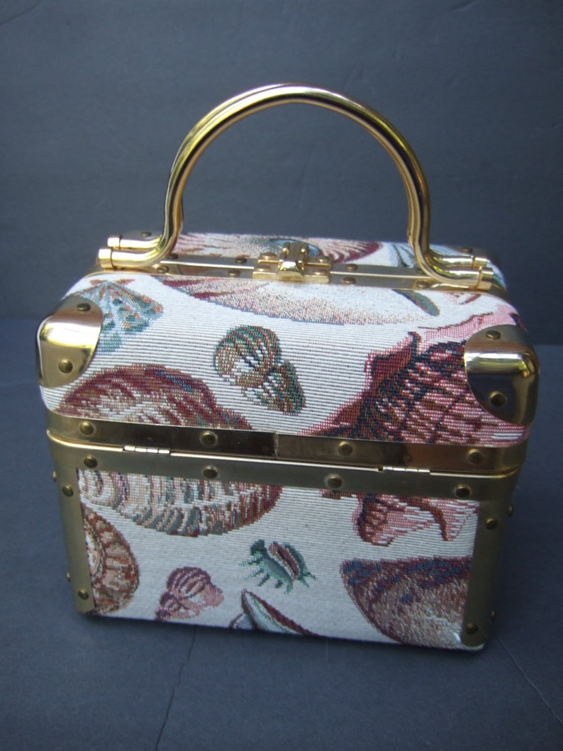 K&ouml;nnte beinhalten: Eine kleine, quadratische Handtasche mit goldenen Verzierungen und einem Tapisserie-Stoff mit Muschelmotiv. Die Tasche hat einen goldenen Henkel und einen Klappverschluss.