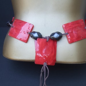 May include: A red and black beaded necklace with square-shaped red beads and black beads. The necklace is tied with a brown string.