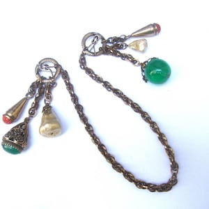 May include: A gold chain necklace with multiple charms, including a pearlescent charm, a green glass bead, and a red and gold charm. The chain is a twisted rope design.