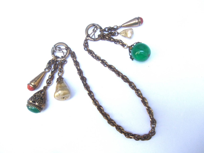 May include: A gold chain necklace with multiple charms, including a pearlescent charm, a green glass bead, and a red and gold charm. The chain is a twisted rope design.