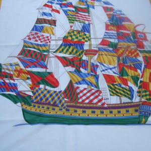 May include: A white fabric with a colorful image of a ship with many flags. The ship is green, yellow, and blue with red, white, and blue flags.