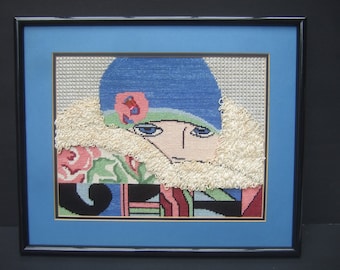 May include: A framed cross-stitch artwork depicting a woman wearing a blue hat with a white fur collar. The woman's face is stylized with large eyes and a pink flower on her hat. The background is a colorful abstract pattern.