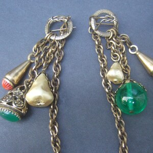 May include: A pair of gold tone earrings with multiple chains and charms. The charms include a pearlescent white pear, a green glass sphere, a red coral teardrop, and a gold tone teardrop with a red coral tip.