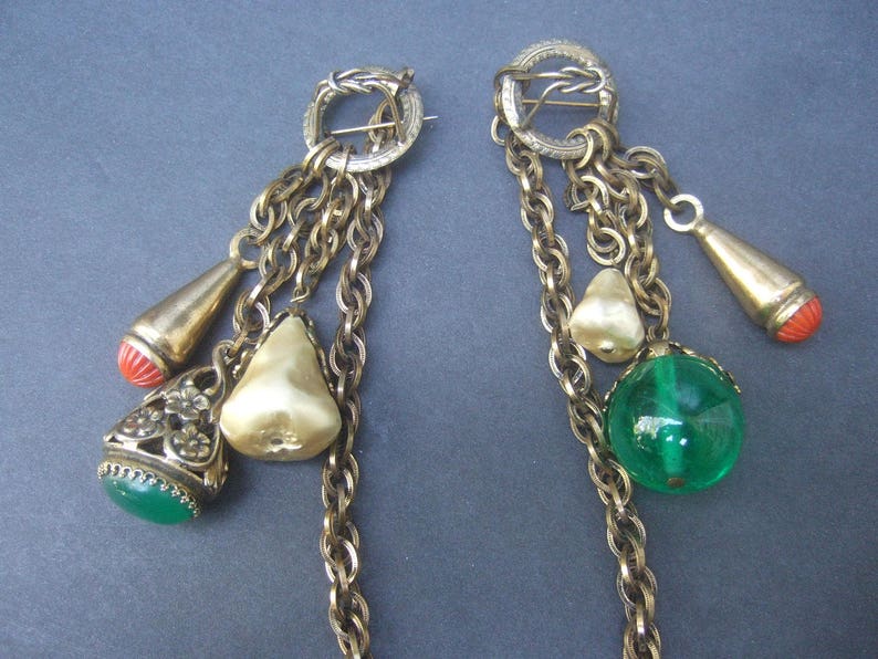 May include: A pair of gold tone earrings with multiple chains and charms. The charms include a pearlescent white pear, a green glass sphere, a red coral teardrop, and a gold tone teardrop with a red coral tip.
