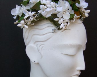 May include: A white floral headpiece with delicate white flowers and green leaves. The headpiece is adorned with a dark green velvet ribbon and is displayed on a white mannequin head against a black background. The flowers appear to be made of fabric.