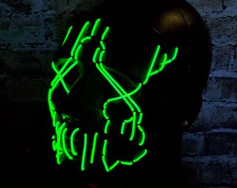 May include: Black mask with bright green neon lines outlining the face. The neon lines create a stylized design across the eyes, nose, and mouth. The mask is set against a dark brick wall.