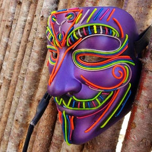 Pode incluir: Uma máscara roxa com um design de rosto sorridente e detalhes em fio de néon colorido. A máscara tem uma alça preta e um cabo preto preso. Os fios de néon são em tons de laranja, amarelo, verde, azul e rosa.