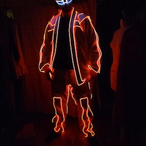Puede incluir: Una persona con un traje naranja brillante con contornos naranja neón y una máscara a juego con detalles azules y blancos. El traje incluye una chaqueta y pantalones, iluminados en un ambiente oscuro.