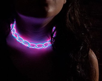 May include: A close-up of a person wearing a glowing pink and blue chain necklace. The necklace is made of plastic and is lit up with LED lights.