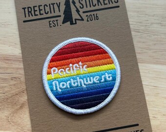 May include: A circular embroidered patch featuring a horizontal rainbow gradient from red to dark blue, with the white text Pacific Northwest centred. The patch is mounted on a brown cardboard display card labelled Treecity Stickers Est. 2016.