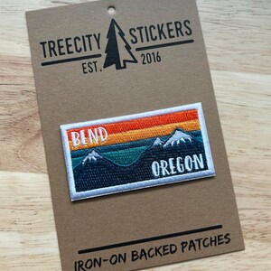 May include: Embroidered patch featuring a mountain landscape with the text "BEND OREGON" against a sunset sky. The patch is rectangular with a white border and is displayed on a brown card with the text "TREECITY STICKERS EST. 2016" and "IRON-ON BACKED PATCHES".