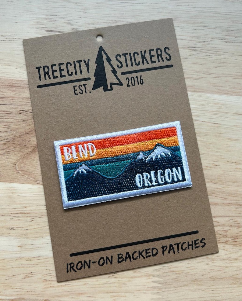 K&ouml;nnte beinhalten: Gestickter Aufn&auml;her mit einer Berglandschaft und dem Text "BEND OREGON" vor einem Sonnenuntergangshimmel. Der Aufn&auml;her hat einen wei&szlig;en Rand und befindet sich auf einer braunen Karte mit "TREECITY STICKERS EST. 2016" und "IRON-ON BACKED PATCHES".